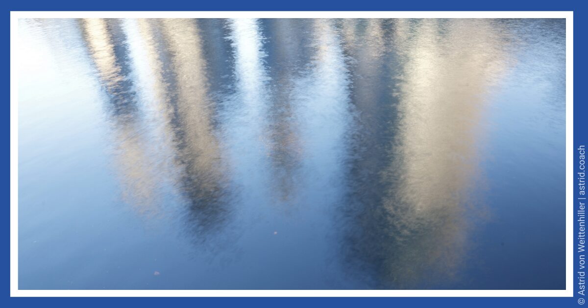 Baumspiegelungen auf ruhiger Wasseroberfläche in Blau- und Goldtönen. Reflections of trees on a calm water surface in shades of blue and gold.